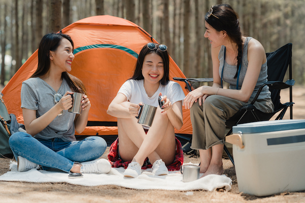 Group of young Asian friends camping or picnic together in forest, teenager female enjoy moment talking in front of their tent. Women do adventure activity and travel on holidays vacation in summer.